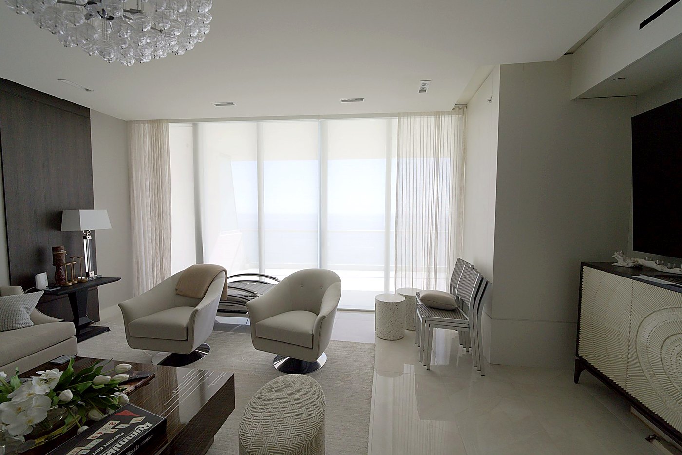 Living room with motorized E-Screen solar shades lowered across the glass wall and sheers at the returns, ocean visible through the screen, Turnberry Ocean Club
