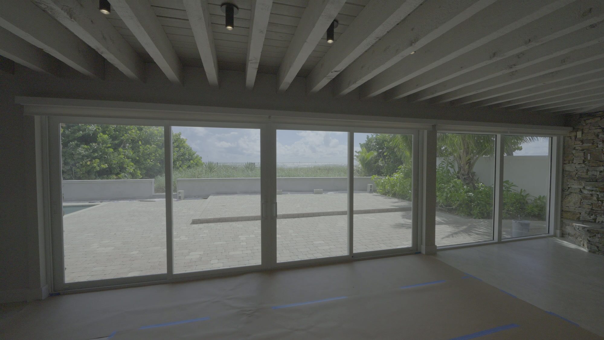 Family room with white beamed ceiling and sliding glass to the pool and sea grapes, roller shades raised