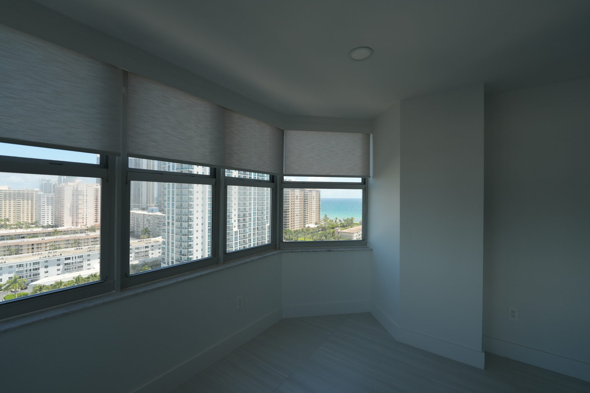 Four roller shades across the city-facing bay windows of a Hallandale Beach condo, with the ocean at the right