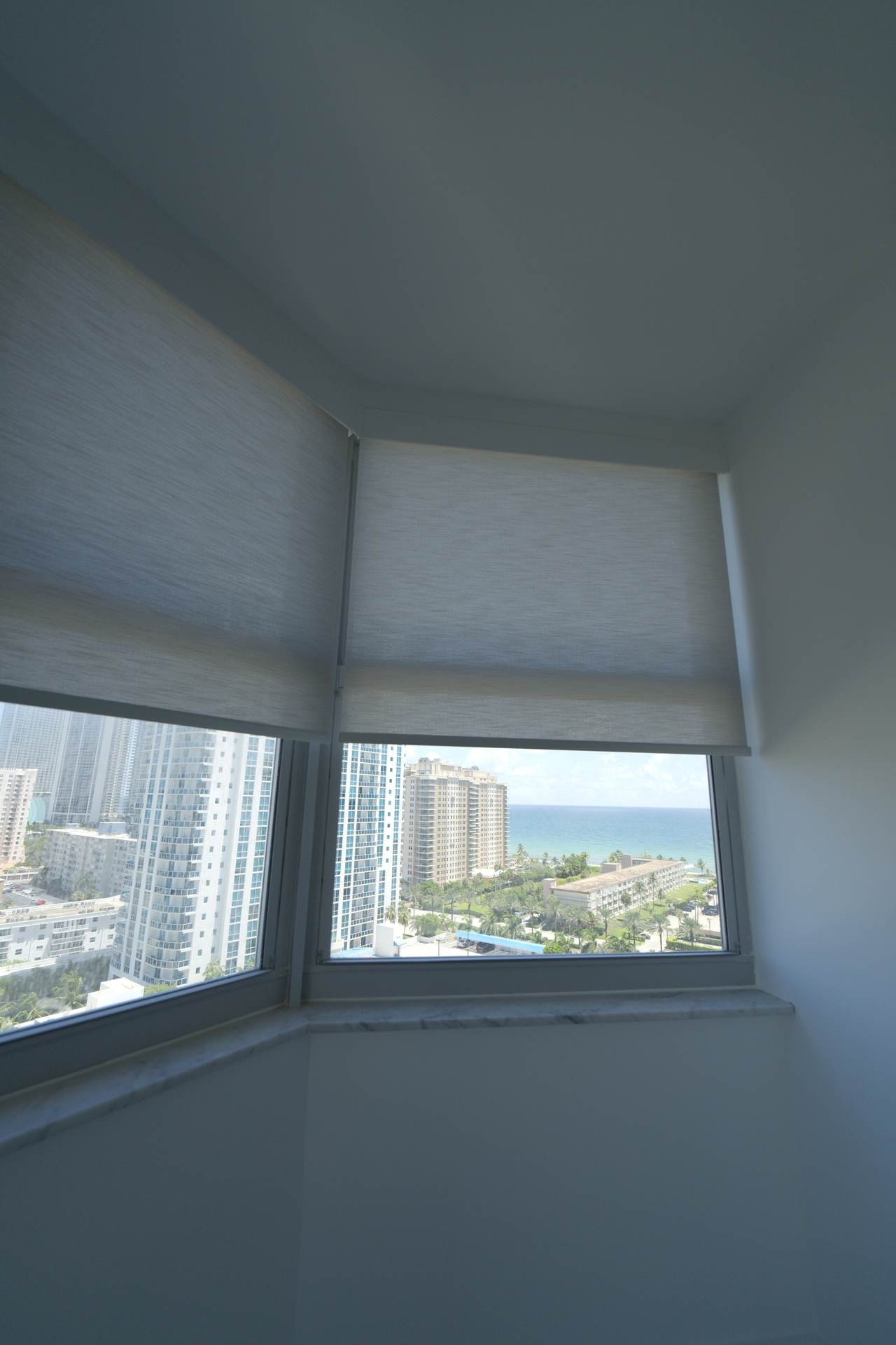 Two roller shades meeting at an angled corner under the lacquered basswood valance at The Hemispheres, Hallandale Beach, halfway down over the ocean view
